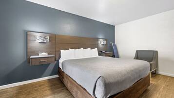 A modern bedroom with a wooden headboard, a grey bedspread, a bedside table with a lamp, and a grey armchair.