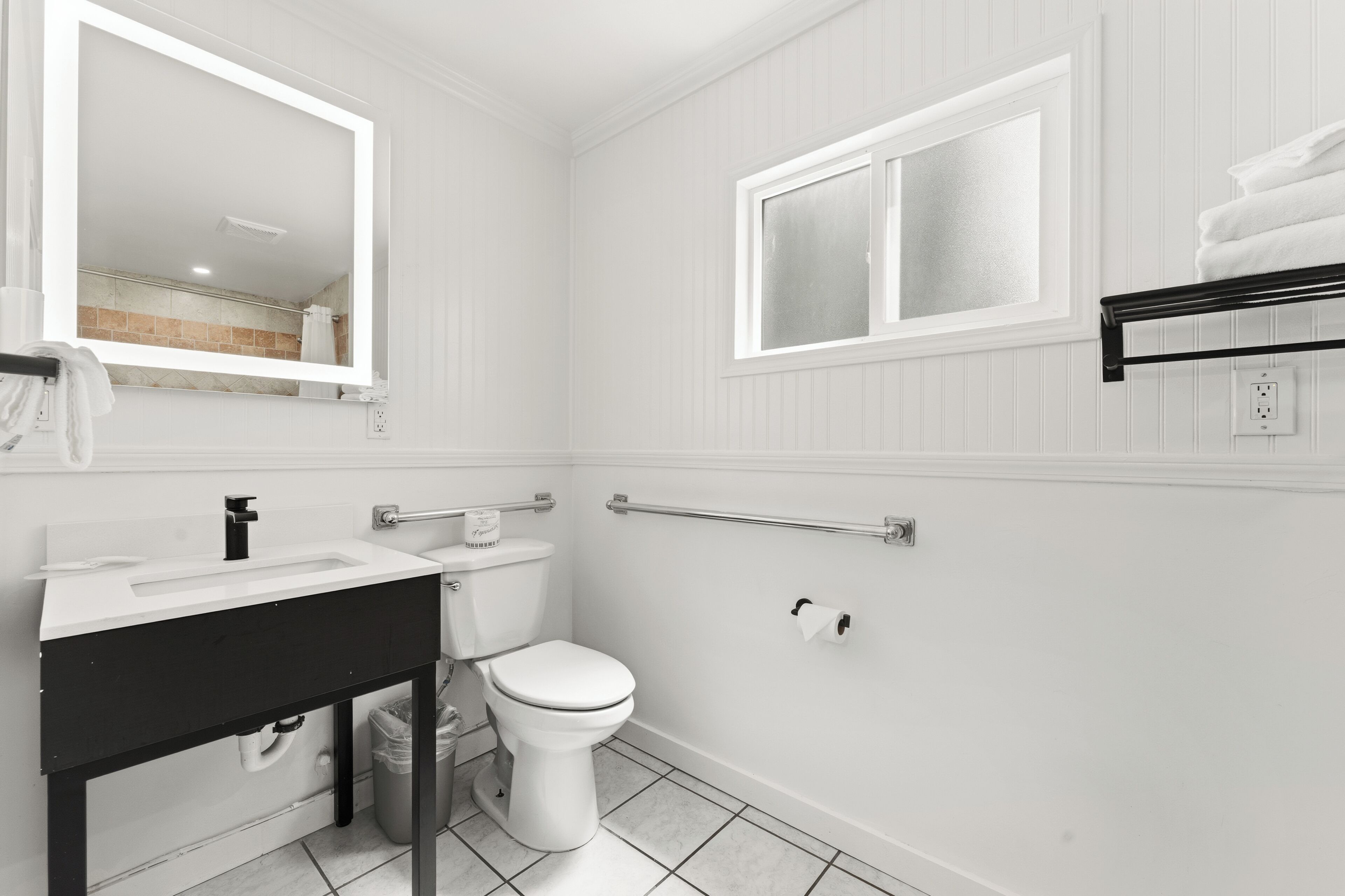 A bathroom with a white sink, toilet, and mirror. There is a window above the toilet and a towel rack on the wall.
