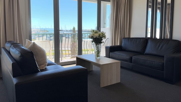 Un salon moderne avec un canapé, une table basse et une vue sur une marina, visible à travers de grandes fenêtres.