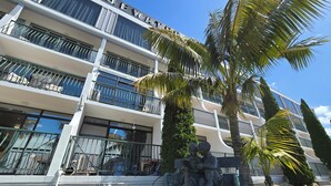 Un hôtel à plusieurs étages doté de balcons, une statue représentant deux personnages en train de lire, et un palmier.