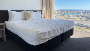 Un grand lit avec une tête de lit sombre et du linge de lit blanc, une table de chevet et une vue sur une marina par une grande fenêtre.