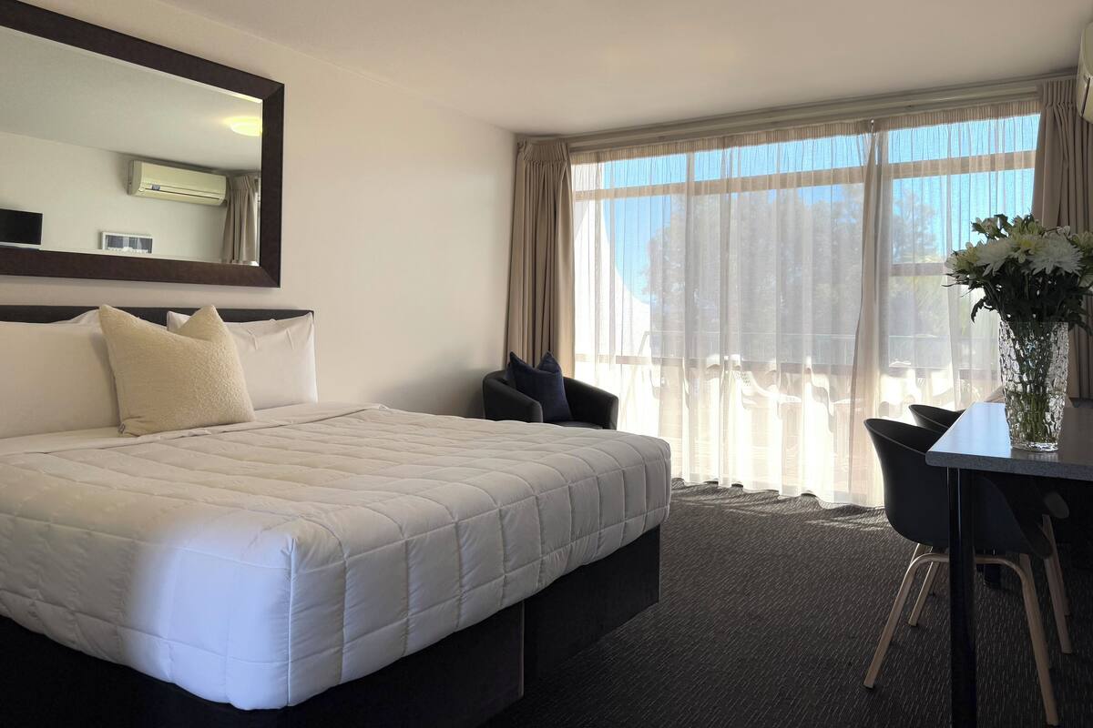 Una habitación de hotel con una cama grande, una silla, una mesa y un florero con flores.