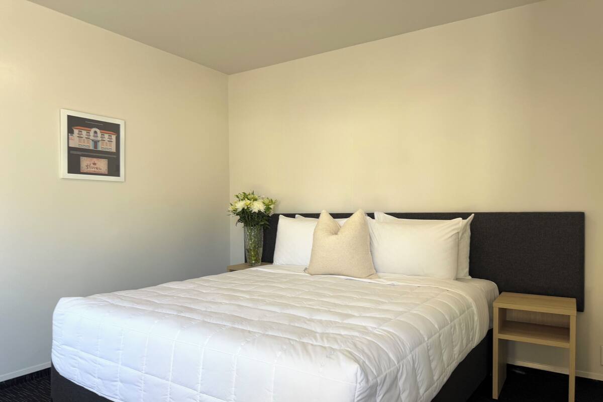 Una cama bien hecha con edredón blanco, una mesita de noche pequeña, un jarrón con flores y un cuadro en la pared.