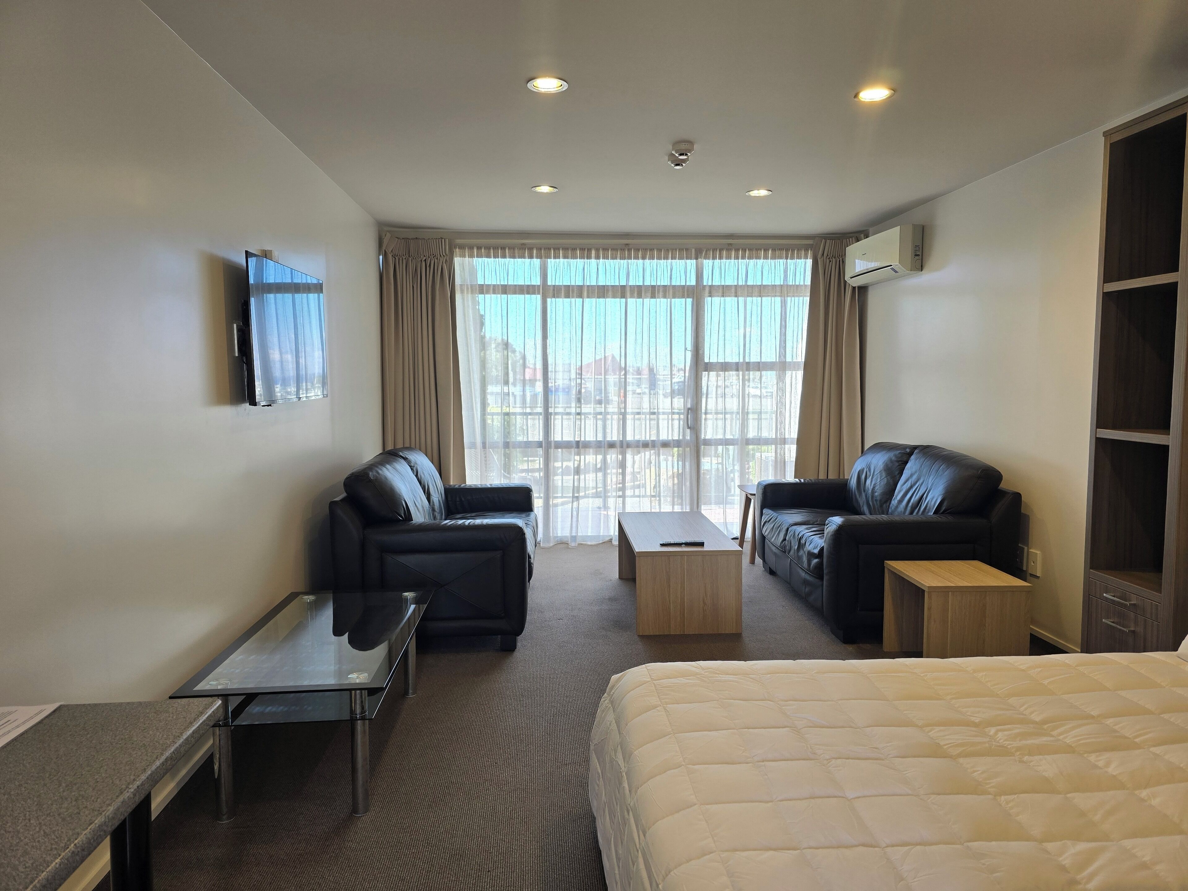 Ein Hotelzimmer mit zwei schwarzen Ledersofas, einem Holztisch, einem Bett mit weißer Daunendecke, einem kleinen Tisch mit Fernbedienung und einem an der Wand befestigten Fernseher.