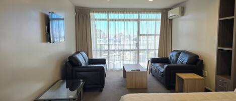 Ein Hotelzimmer mit zwei schwarzen Ledersofas, einem Holztisch, einem Bett mit weißer Daunendecke, einem kleinen Tisch mit Fernbedienung und einem an der Wand befestigten Fernseher.