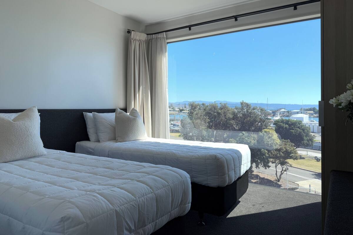 Habitación de hotel con dos camas, un ventanal con vistas a un puerto deportivo y montañas, y una cortina en la ventana.