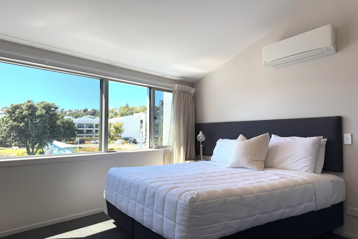 Una habitación de hotel con una cama grande, un ventanal con vistas a edificios y un árbol, y un equipo de aire acondicionado en la pared.