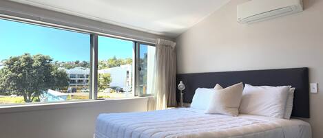 Una habitación de hotel con una cama grande, un ventanal con vistas a edificios y un árbol, y un equipo de aire acondicionado en la pared.