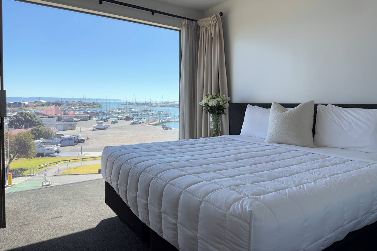 Habitación de hotel con una cama grande, vista a un puerto deportivo y una ventana con cortinas.
