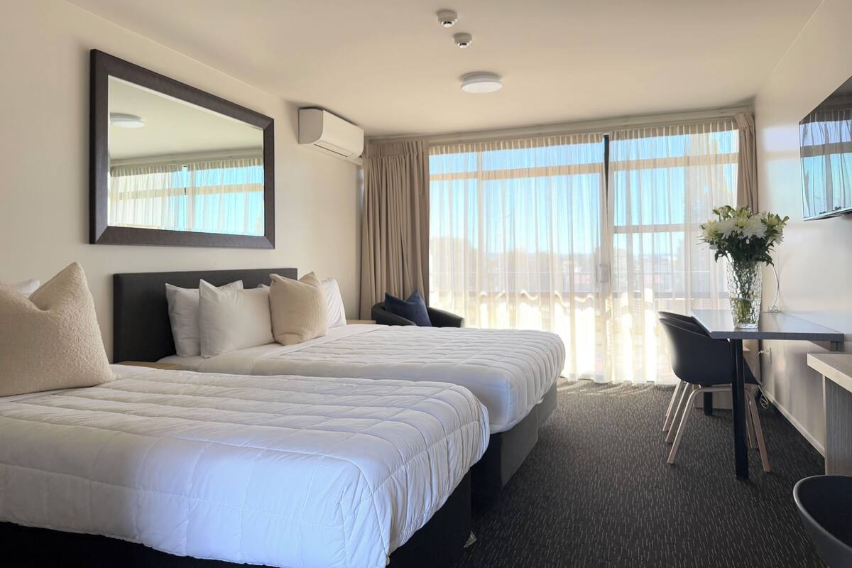 Habitación de hotel con dos camas, un escritorio, una silla, un ventanal con cortinas y un espejo en la pared.