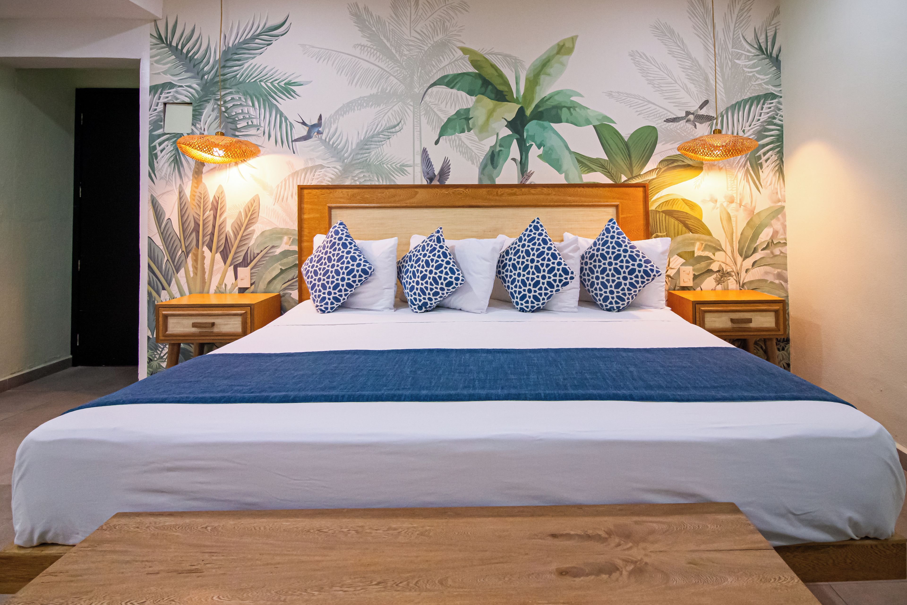 A neatly made bed with a blue bedspread and patterned pillows, a wooden headboard, and a tropical mural on the wall.