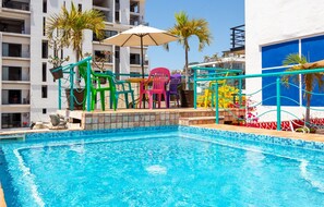 Una piscina con sillas de colores y una sombrilla, rodeada de palmeras y un edificio con balcones.