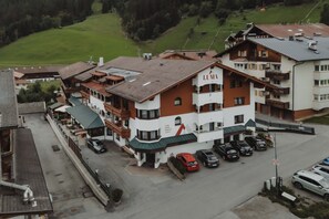 A multi-story hotel with a prominent sign, a parking lot with several cars, and a hilly landscape in the background. - LUMA Stubai Aparthotel (Neustift Im Stubaital)