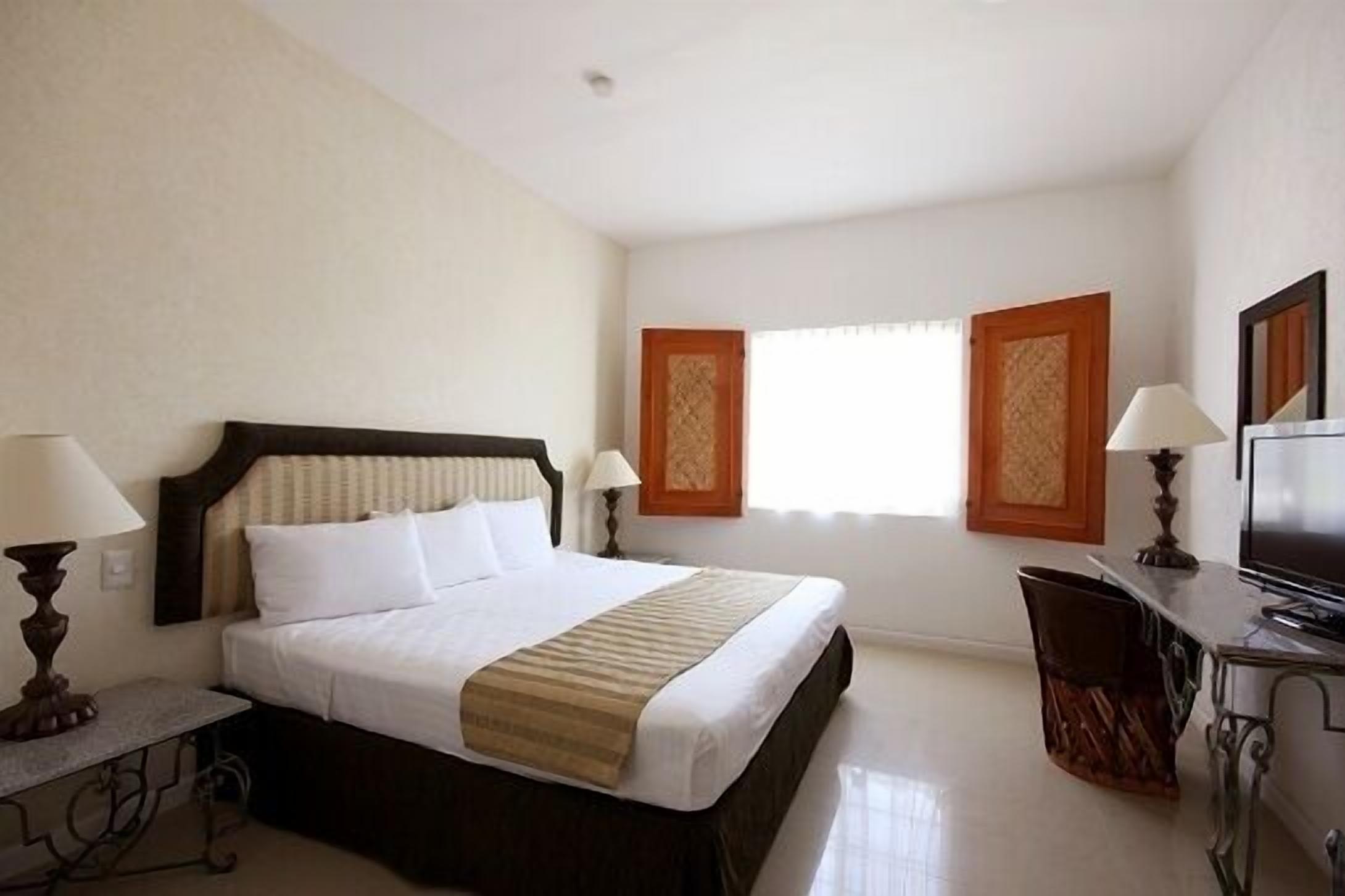 A hotel room with a large bed, a desk, a chair, and two windows with curtains.