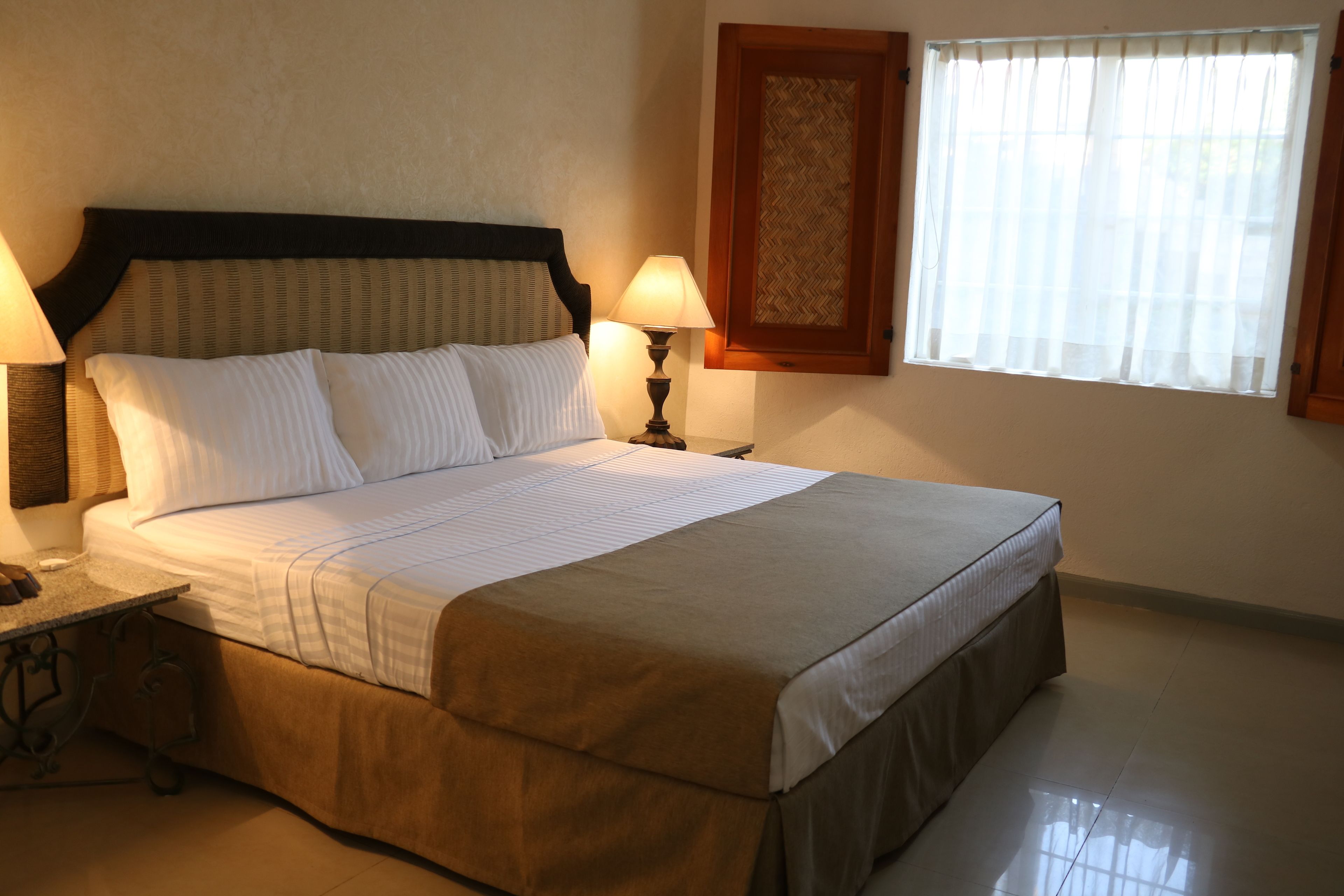 A neatly made bed with a headboard, two bedside lamps, and a window with sheer curtains.