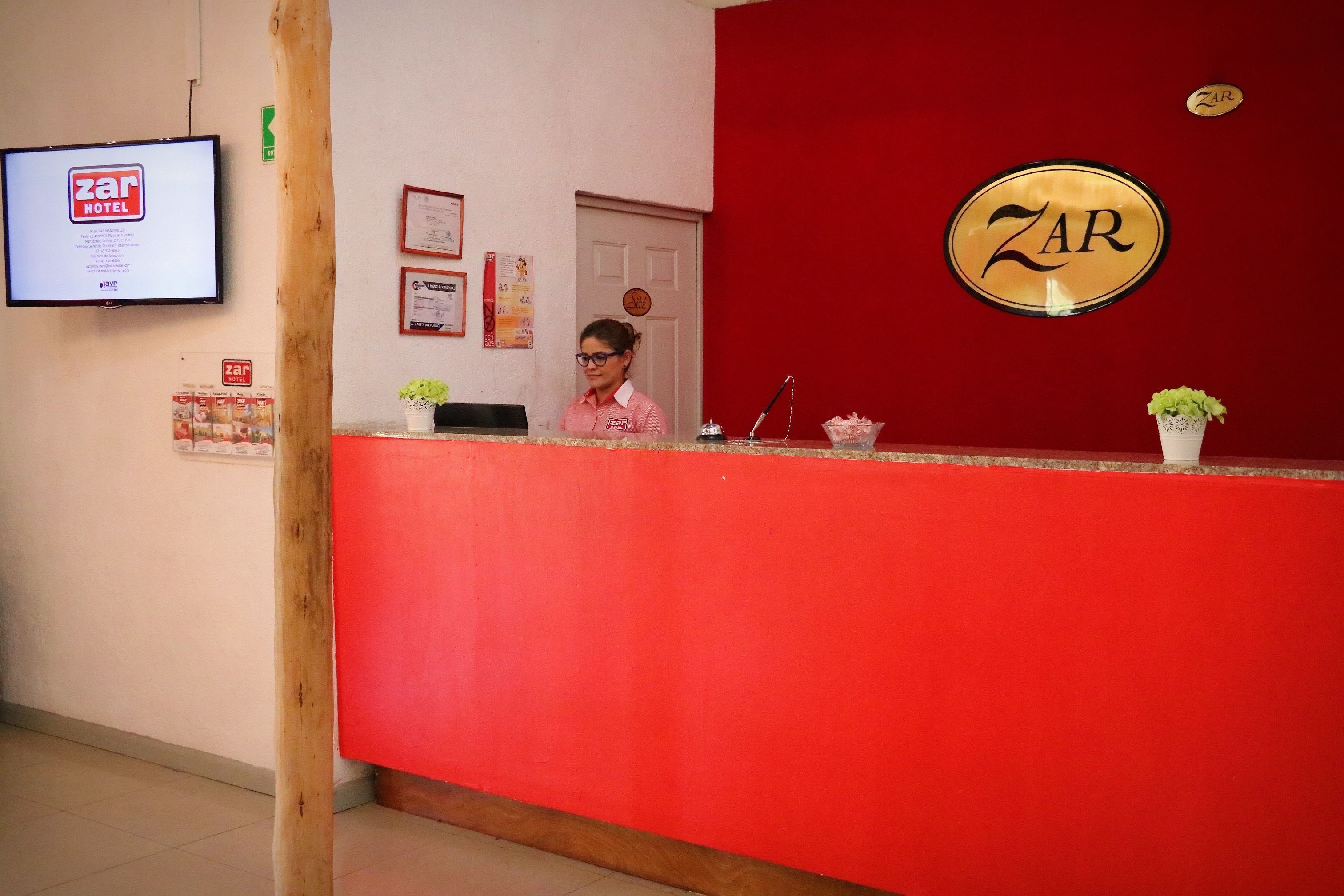 Taulell de recepció d'hotel amb una paret vermella, un senyalitzador 'ZAR HOTEL' i un recepcionista.