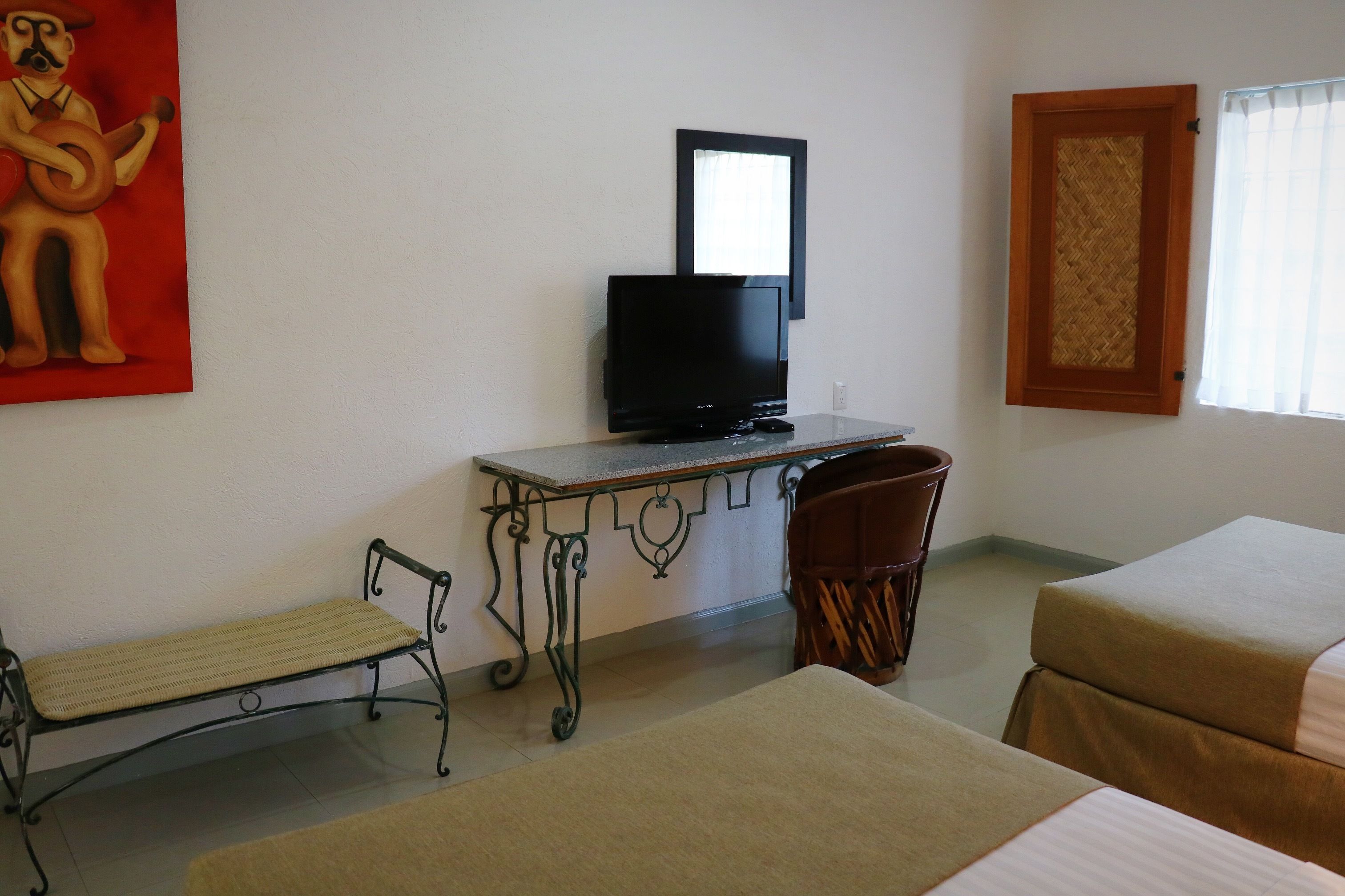 A hotel room with a bed, a desk with a television, a small bench, and a window with a curtain.