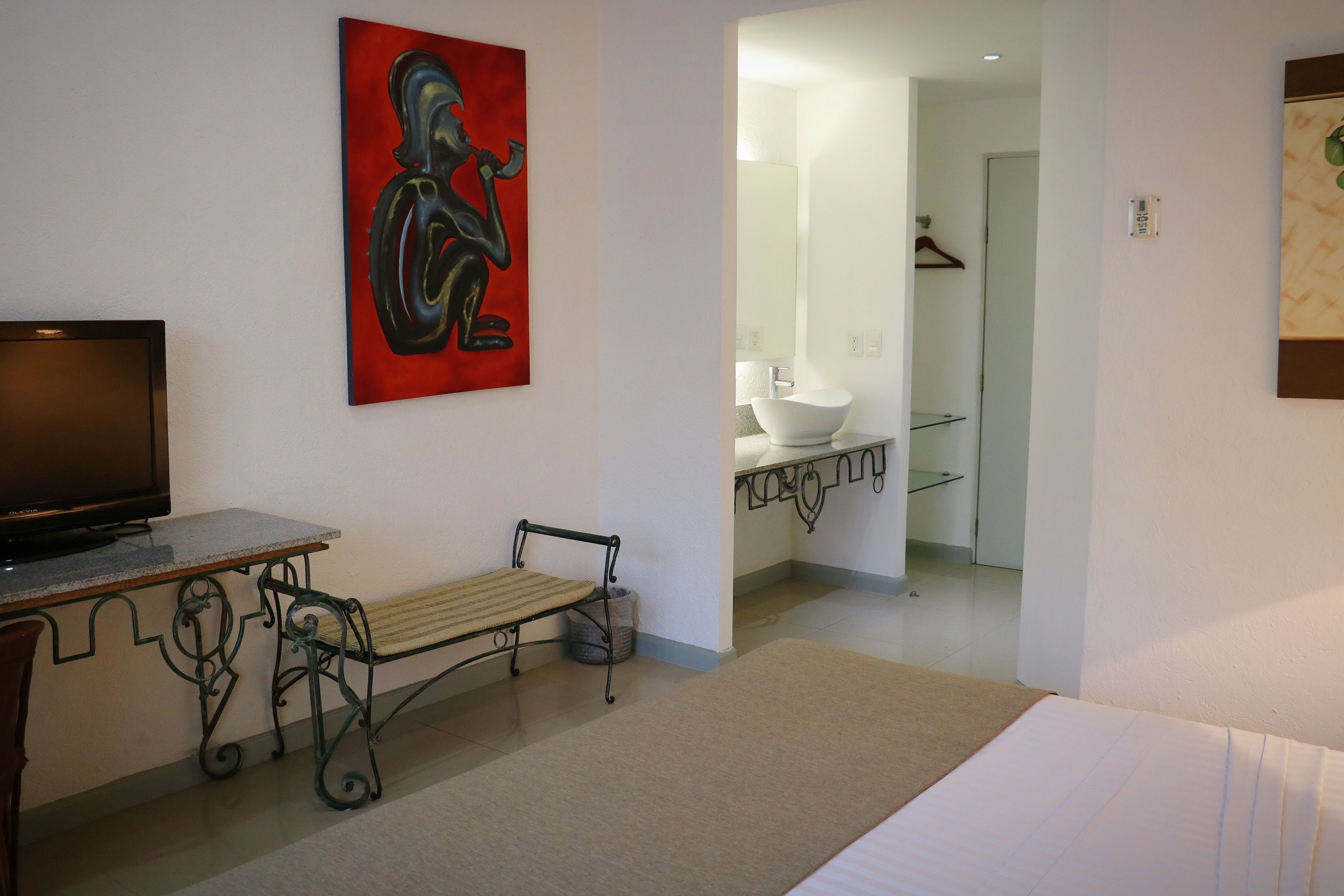 A hotel room with a bed, a TV, a bench, a painting, a bathroom with a sink and mirror, and a hallway.