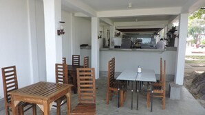 Uma área de refeições ao ar livre coberta, com mesas e cadeiras de madeira, uma mesa branca no centro e uma cozinha ao fundo.