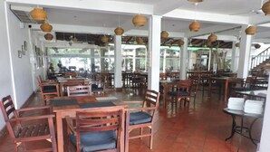 Um restaurante espaçoso com mesas e cadeiras de madeira, luminárias de vime penduradas e amplas janelas que permitem a entrada de luz natural.