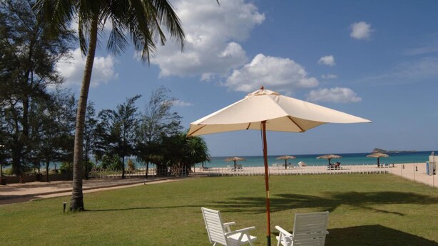 Uma área gramada com duas cadeiras de praia brancas e um guarda-sol bege, com vista para uma praia com guarda-sóis e palmeiras.