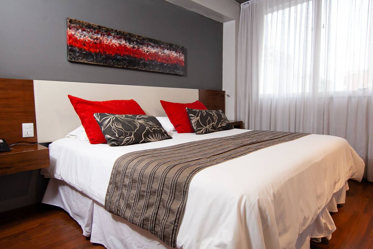 Habitación de hotel con cama, mesita de noche, un cuadro en la pared y una ventana con cortinas.