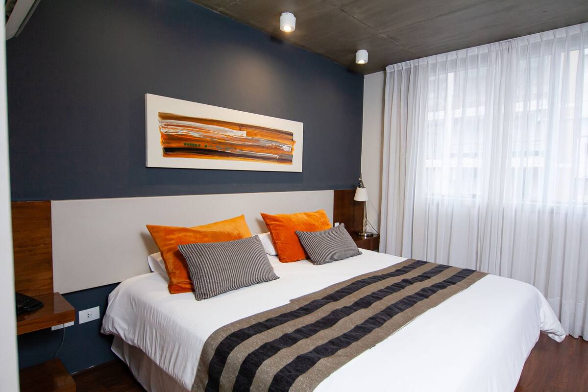 Una cama bien hecha con una colcha a rayas, dos almohadas naranjas y una almohada gris.