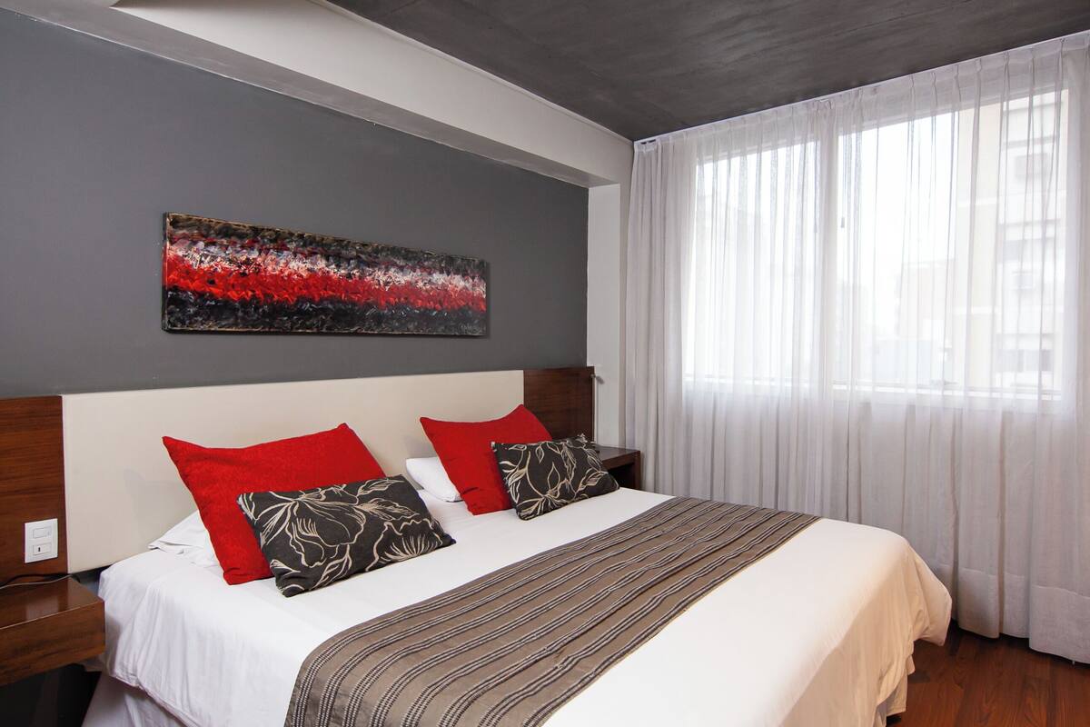 Habitación de hotel con una cama grande, dos almohadas rojas, una almohada estampada, una manta a rayas y un cuadro abstracto moderno en la pared.