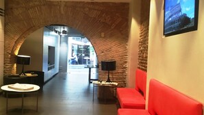 A lobby with a brick archway, a red sofa, a table with a lamp, and a TV displaying a building. - Hotel De Rome (Rome)