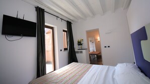 A bedroom with a bed, a flat-screen TV, a window with curtains, and a hallway leading to a bathroom. - Hotel De Rome (Rome)