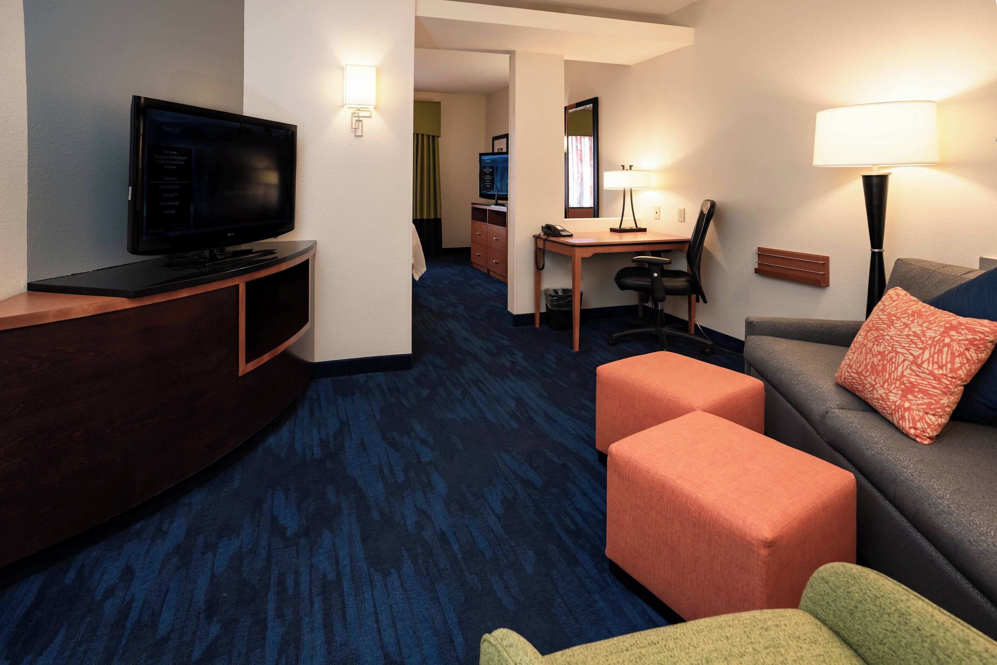 A hotel room with a flat-screen TV, a desk with a chair, a sofa with ottomans, and a lamp.