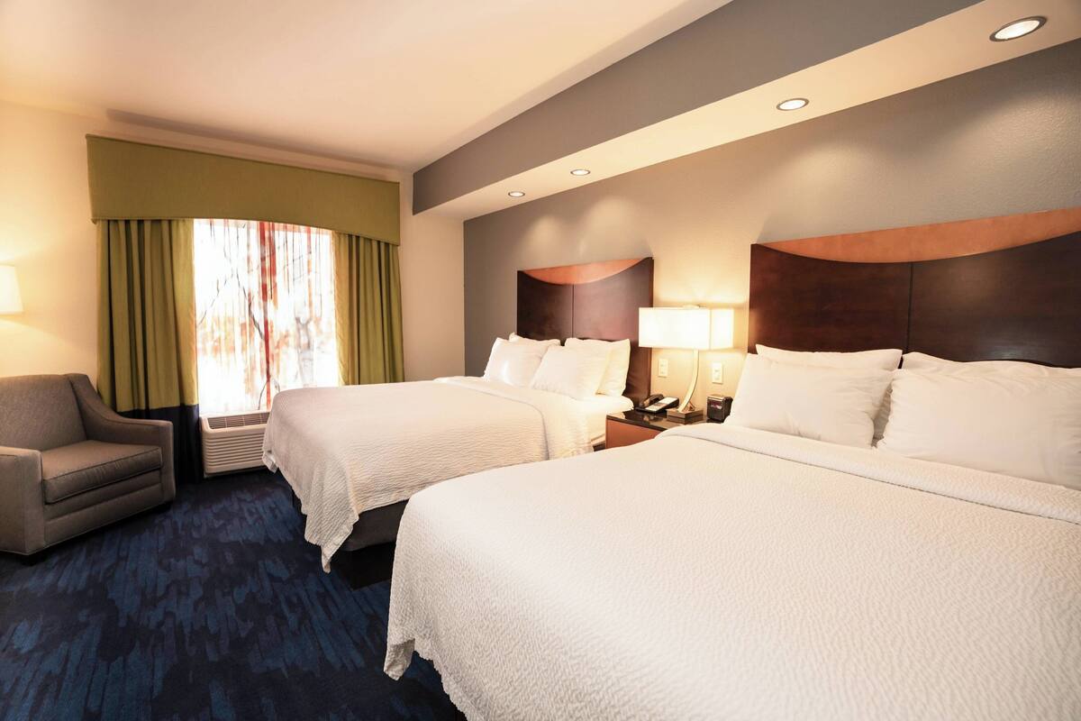 A hotel room with two beds, a chair, a lamp, and a window with curtains.