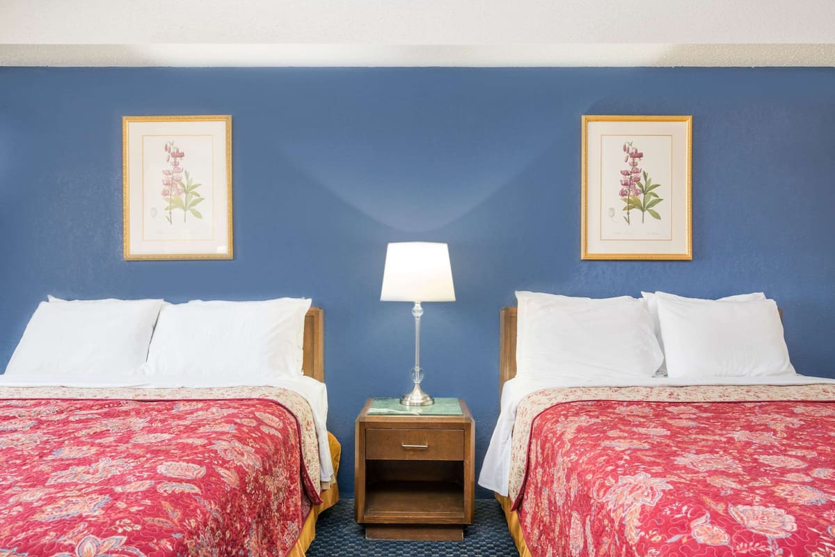 A hotel room with two beds, a nightstand with a lamp, and two framed botanical prints on the wall.