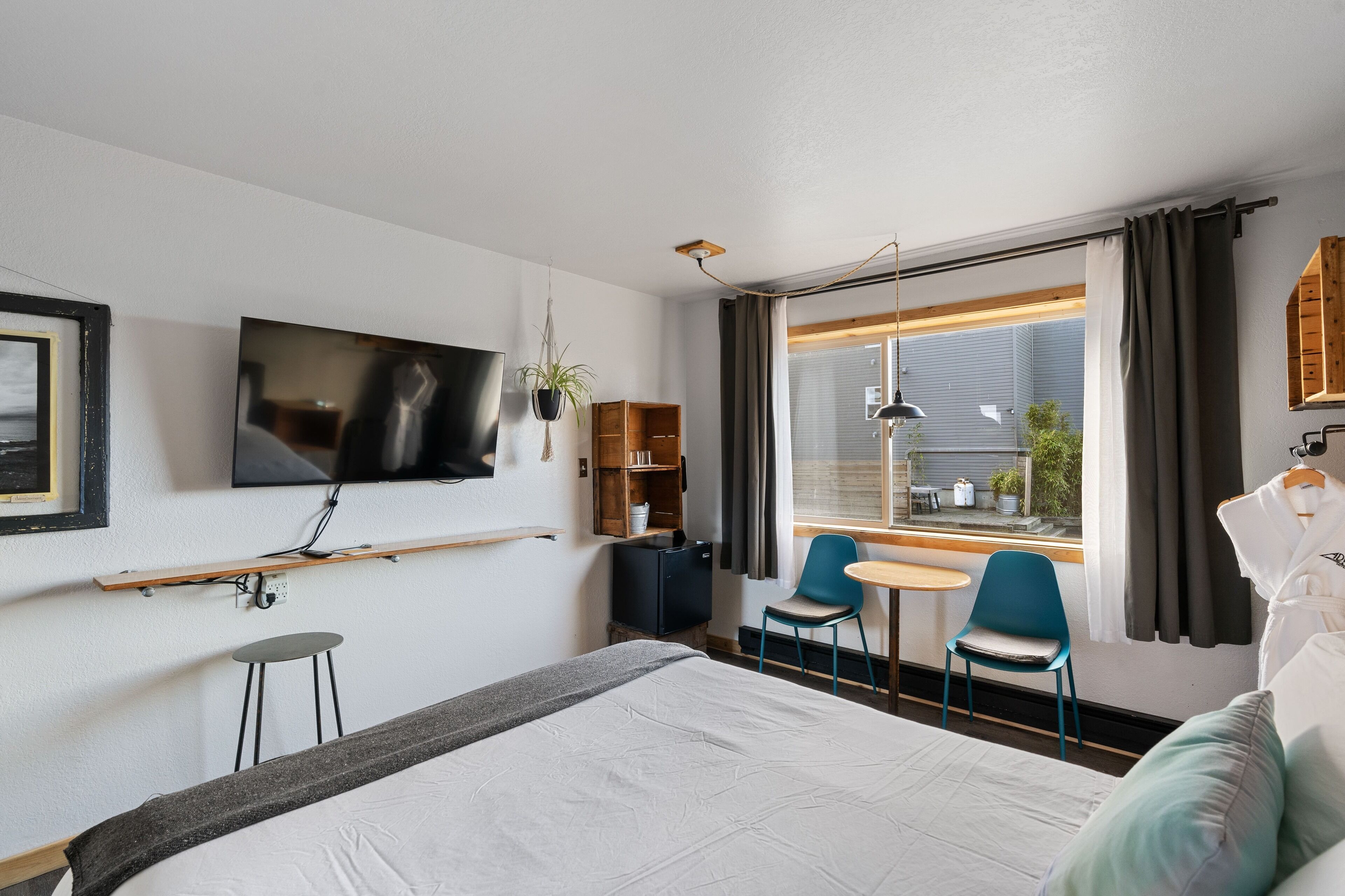 Una habitación de hotel con una cama, un televisor montado en la pared, una pequeña mesa con dos sillas, un escritorio, un armario y una ventana con cortinas.