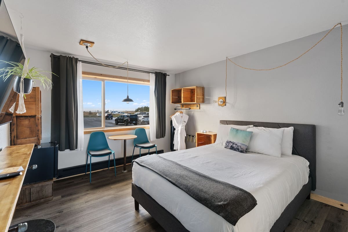 Ein Hotelzimmer mit einem großen Bett, einem Schreibtisch, einem Sessel, einem kleinen Tisch, einer Zimmerpflanze und Blick ins Freie.