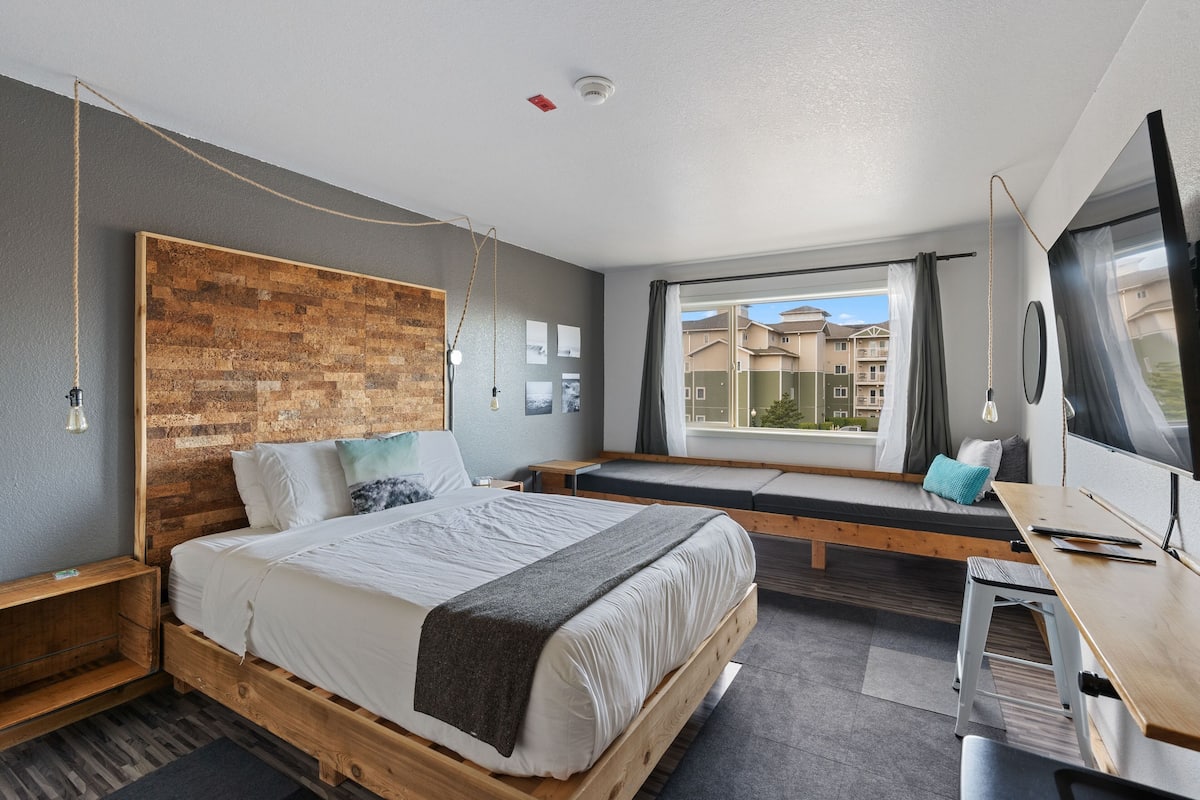 Ein modernes Schlafzimmer mit einem Holzkopfende, einem großen Bett, einem Schreibtisch und einem Fenster mit Blick auf Gebäude.