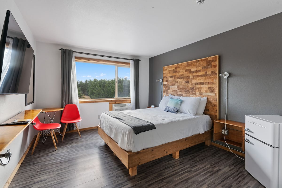 Ein modernes Schlafzimmer mit einem hölzernen Bett, einem Schreibtisch, roten Stühlen, einem Fernseher und einem Fenster mit Blick auf Bäume.
