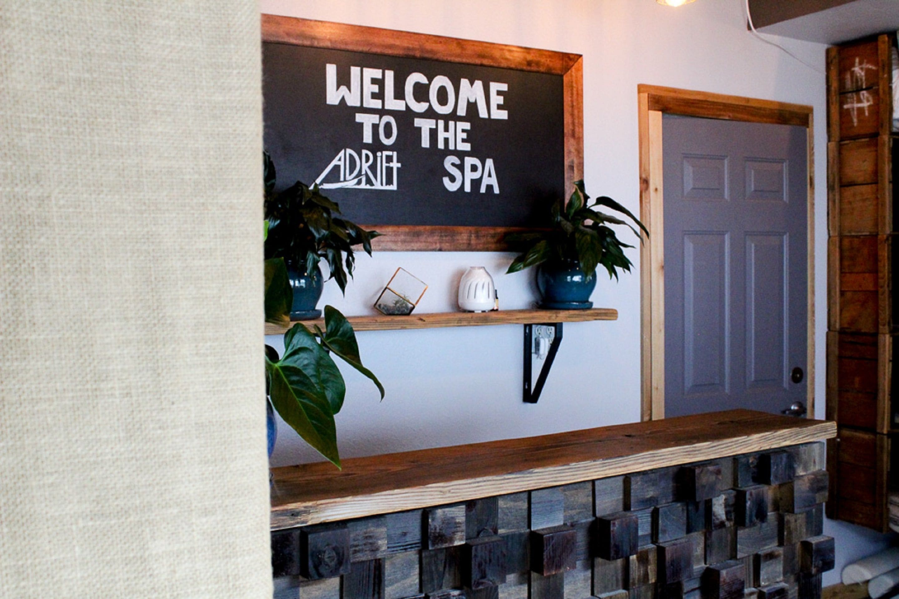 A reception area with a welcome sign, two potted plants, a small shelf with decorative items, and a wooden bench.