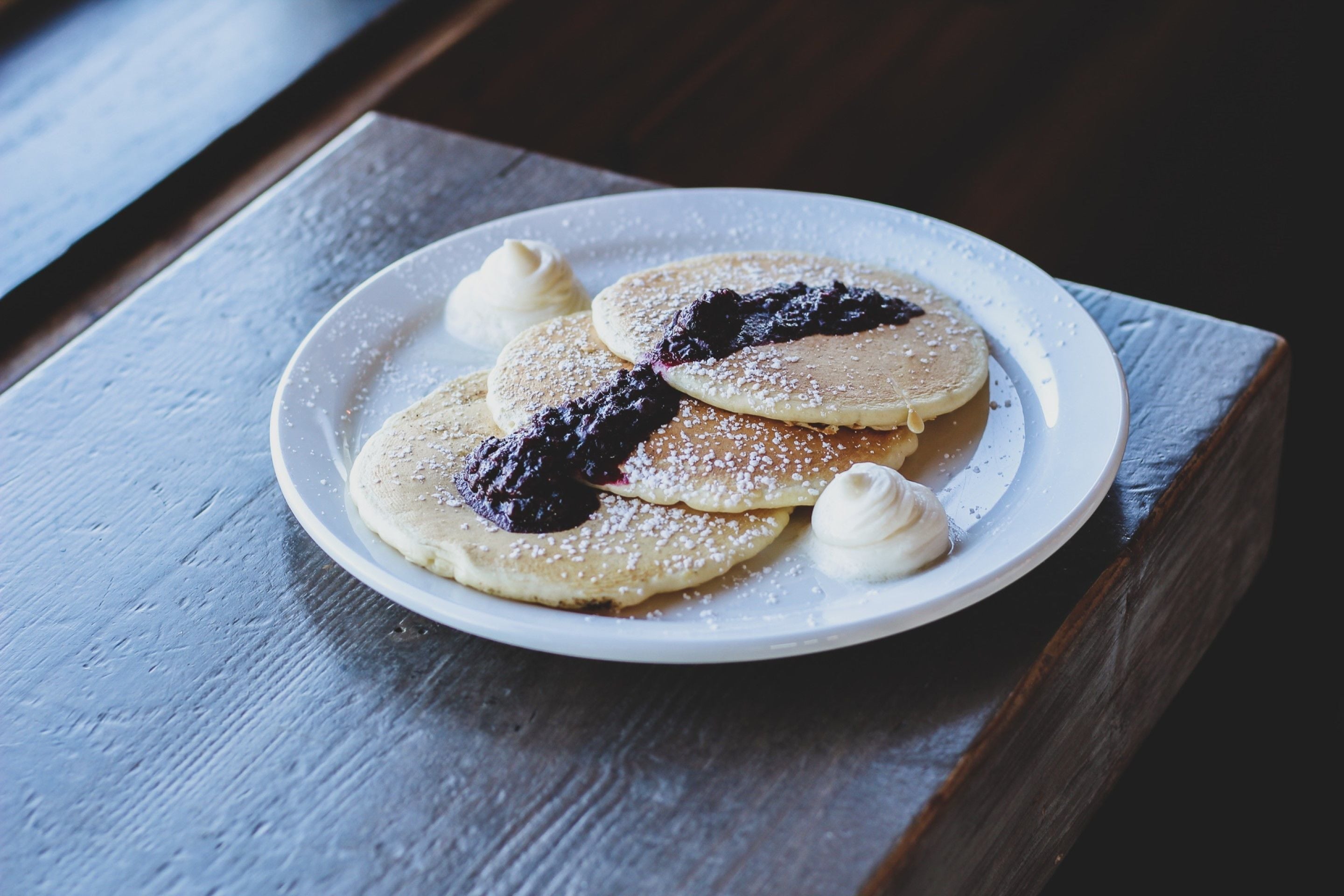 Een bord pannenkoeken met blauwe bessencompote en slagroom op een houten tafel.
