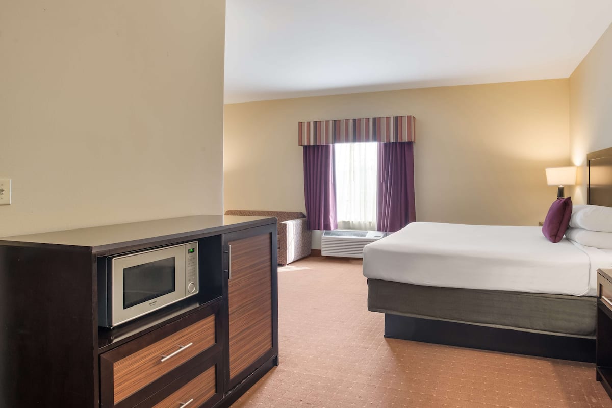 A hotel room with a bed, a desk with a microwave, a chair, and a window with curtains.