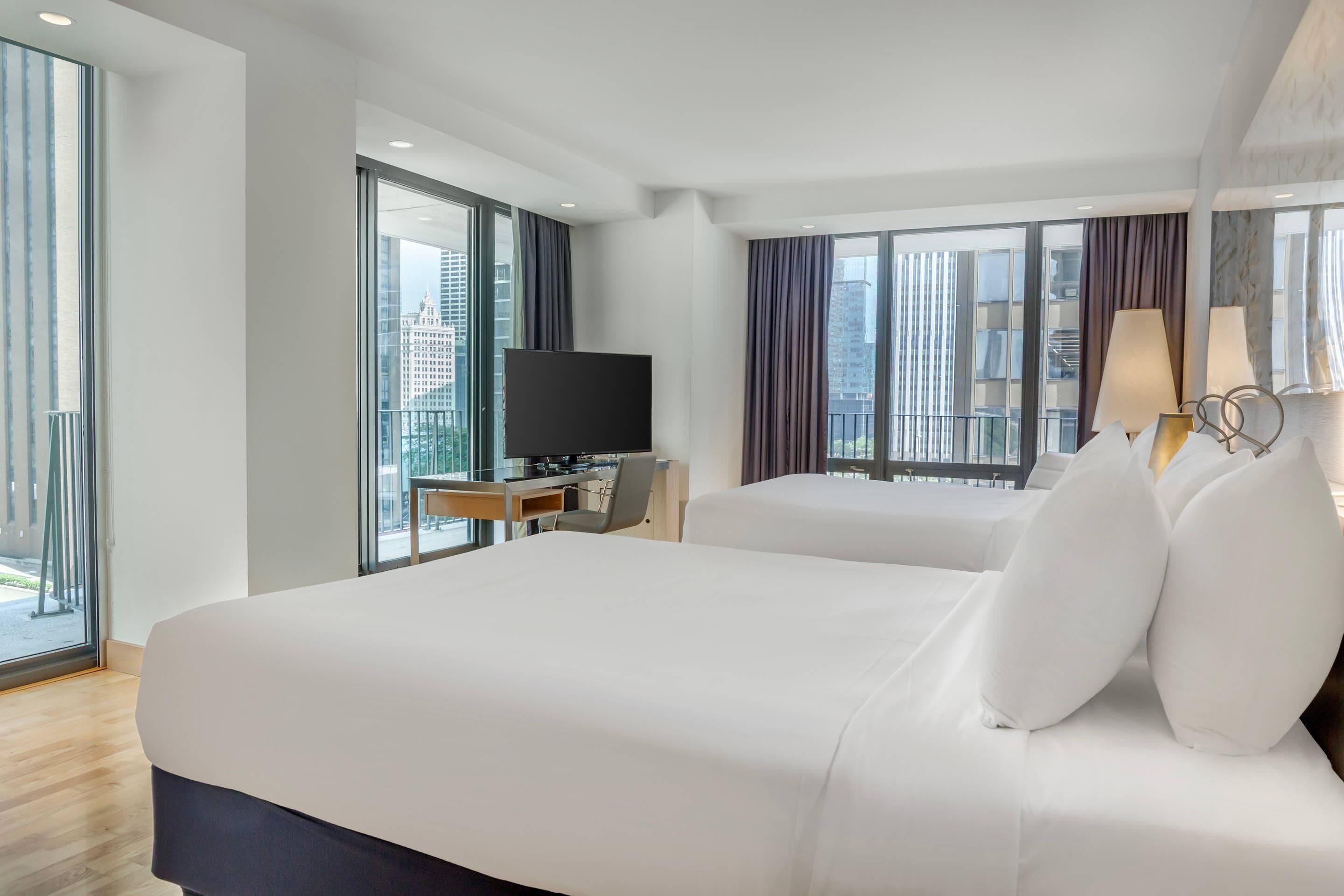 A hotel room with a large bed, a desk with a television, and a view of the city through large windows.