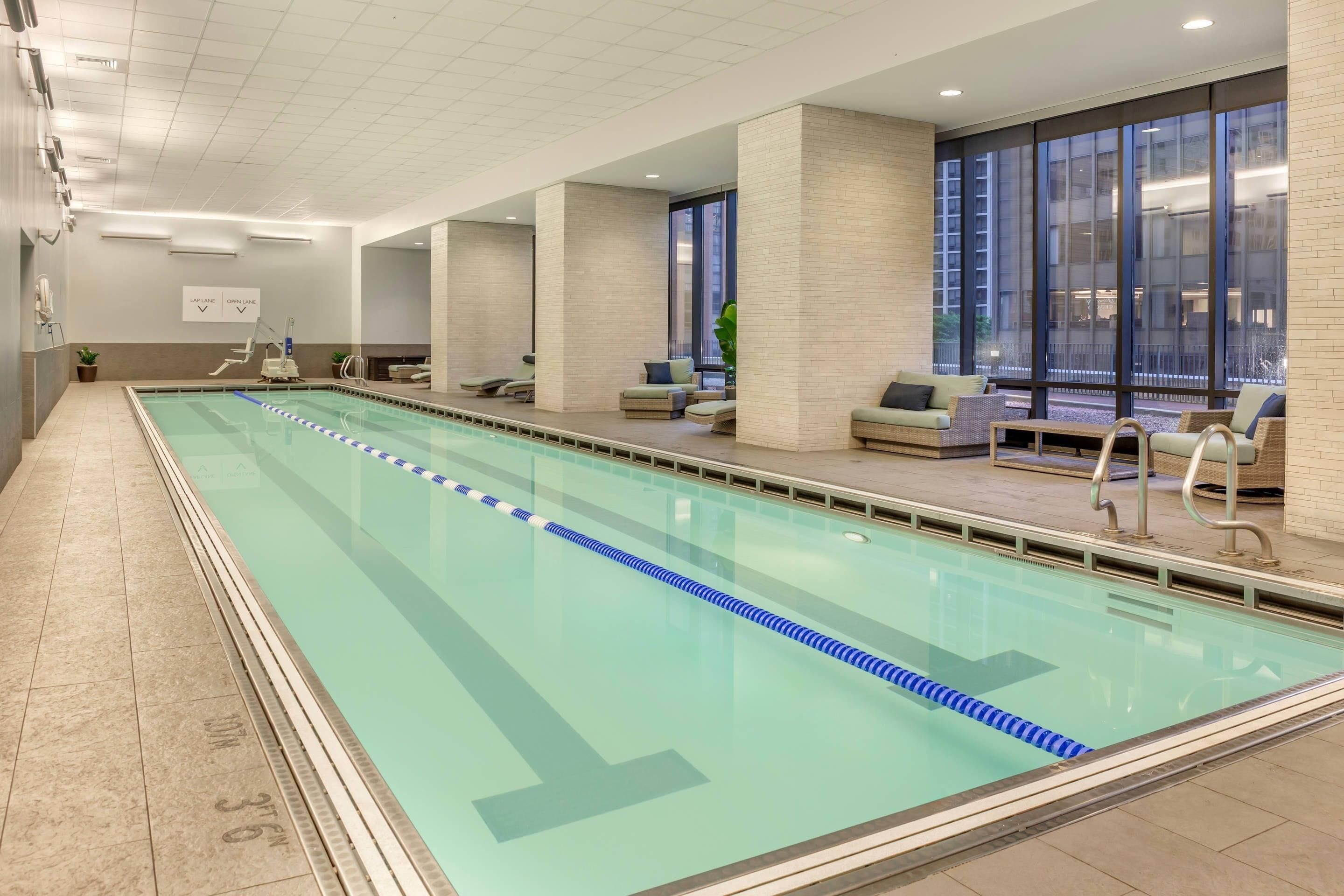 A long indoor swimming pool with clear lane dividers, a tiled floor, and a seating area with sofas and chairs.