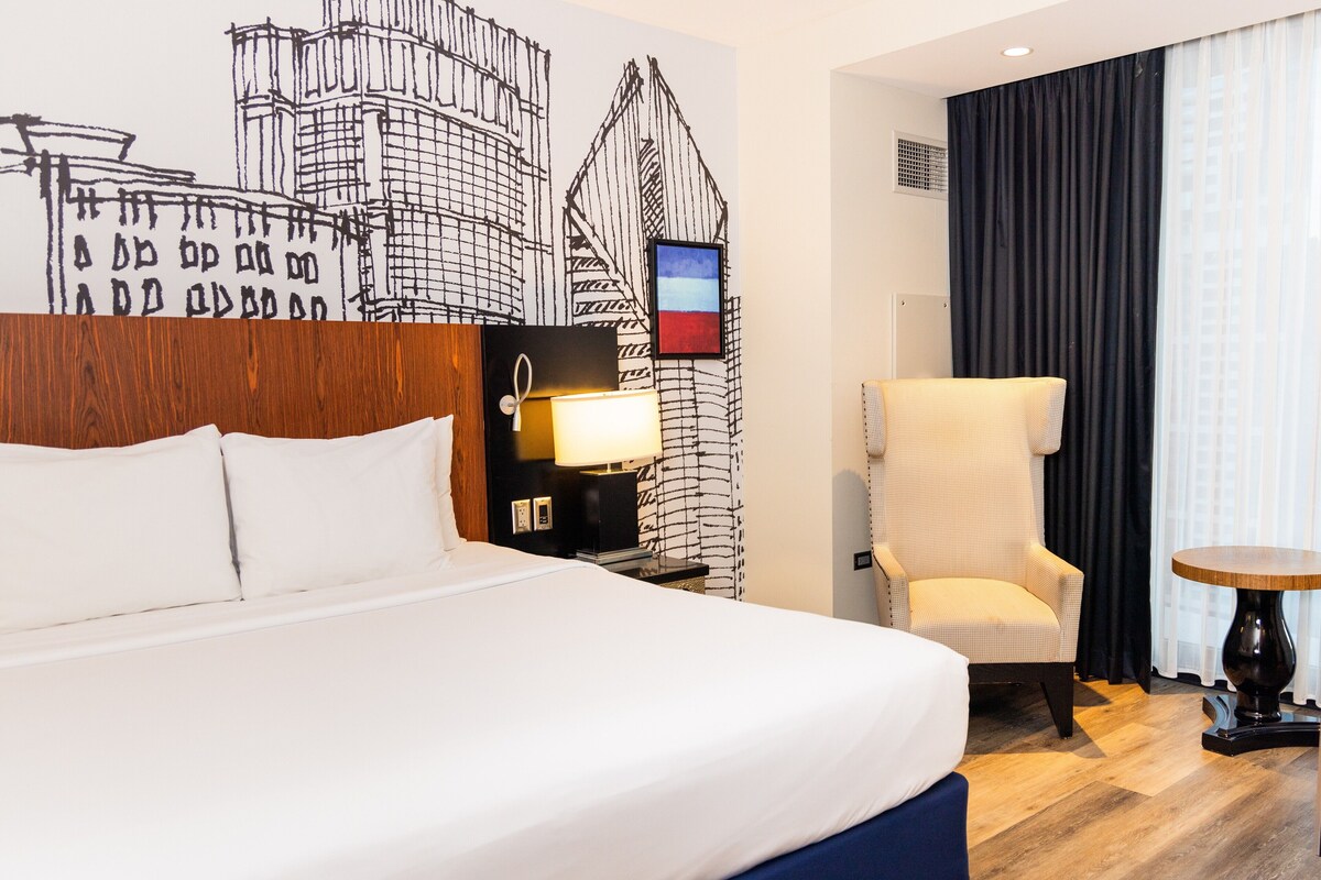 A hotel room with a large bed, a chair, a small table, and a wall with a black and white abstract design.