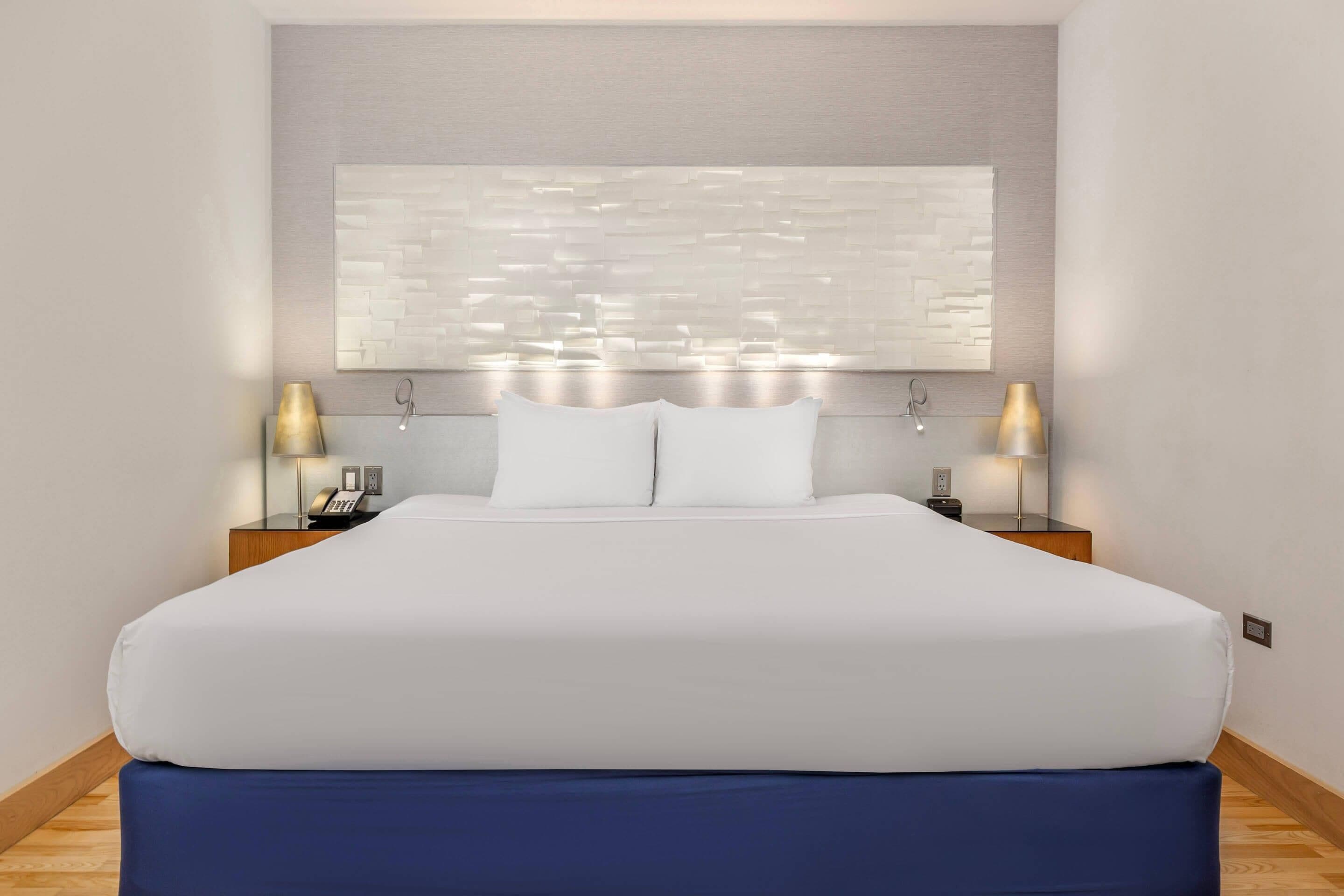 Una habitación de hotel con una cama grande, dos mesitas de noche, una lámpara de pared y una obra de arte abstracta moderna.