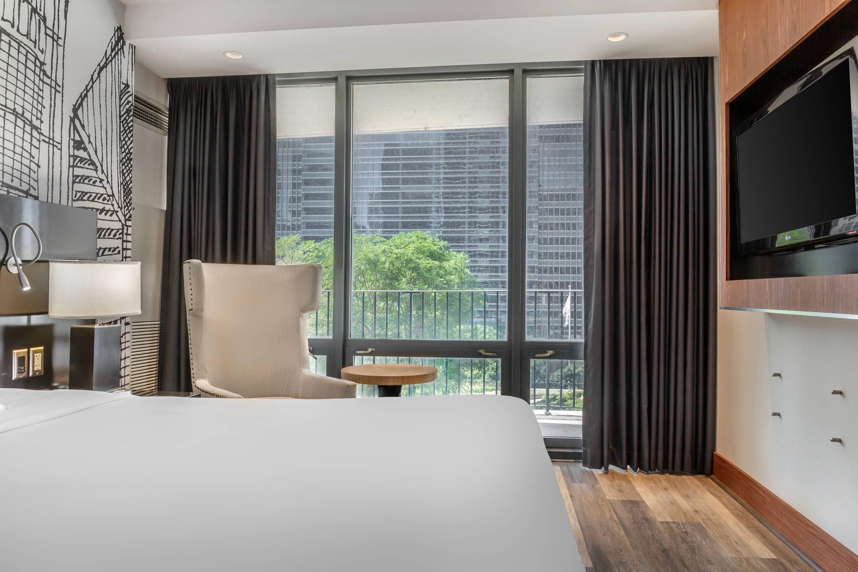 Habitación de hotel con una cama grande, una silla, una mesita, televisor y un balcón con vistas a edificios y árboles.