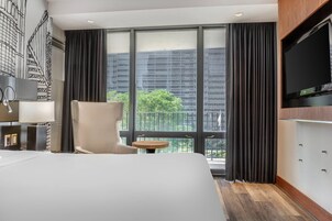 A hotel room with a large bed, a chair, a small table, a TV, and a balcony with a view of buildings and trees.