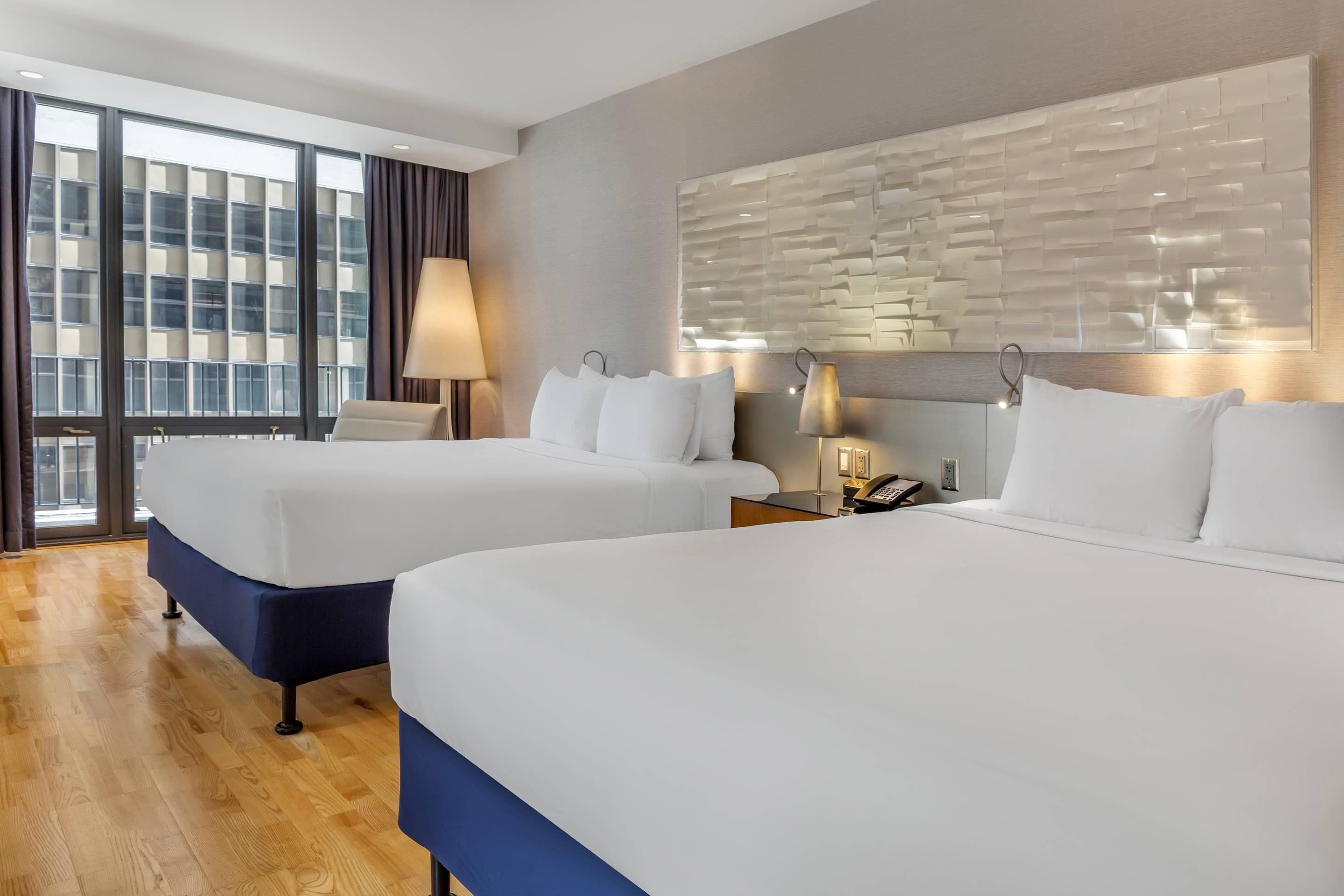 A hotel room with two beds, a large window with curtains, a wall-mounted lamp, and a modern wall design.
