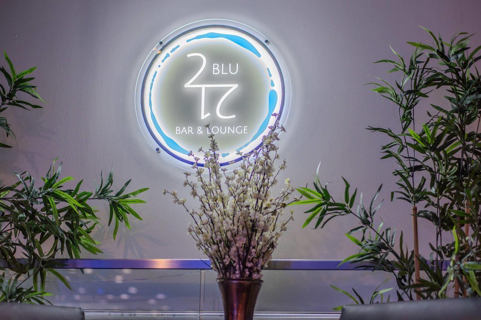 En rund neonskylt med texten ”BLU 7 BAR & LOUNGE”, omgiven av krukväxter.