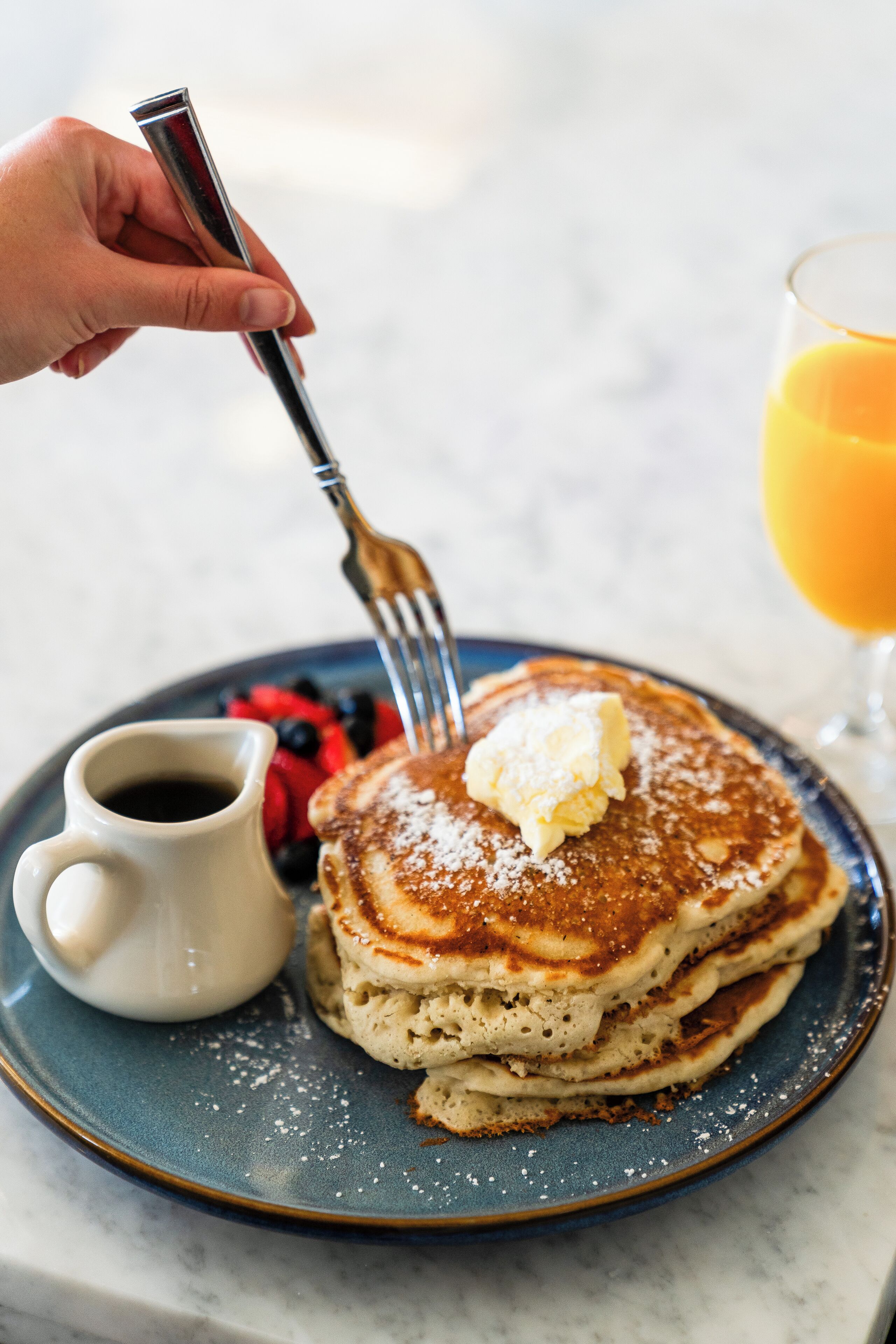 A stack of pancakes with butter and powdered sugar, a fork, a glass of orange juice, and a cup of coffee on a plate.