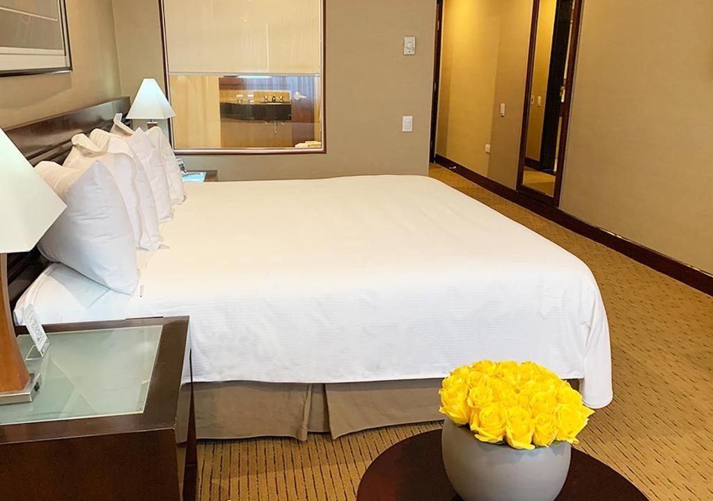 Ein Hotelzimmer mit einem großen Bett, einem Nachttisch mit Lampe und einer Vase mit gelben Blumen.